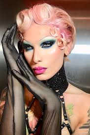loseup portrait of a drag queen with vibrant pink hair styled in victory rolls, adorned with delicate pink flowers. she's wearing dramatic eye makeup with a blend of blues and greens, and her lips are painted a bold pink.  she's accessorized with long black lace gloves and a sparkling black choker. her expression is confident and alluring. the background is blurred, drawing focus to the queen's striking features and costume. the image exudes glamour and theatrical flair.