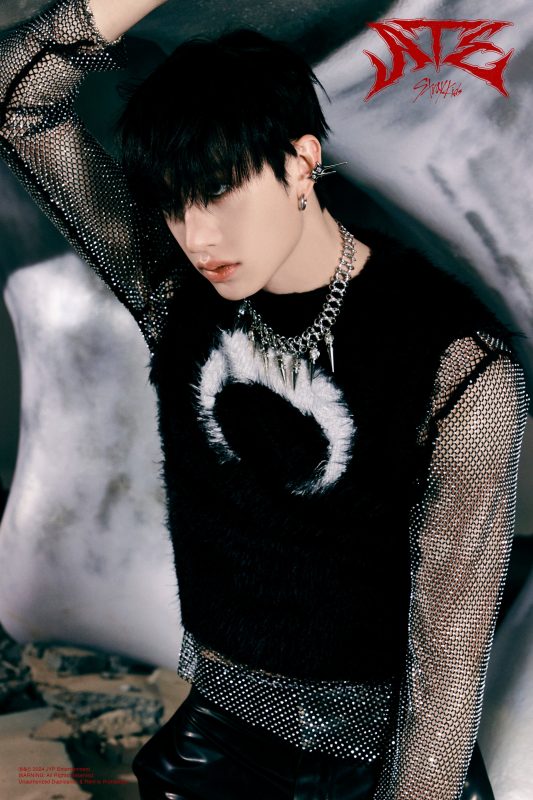 a closeup portrait of stray kids' i.n (jeong in) from a photoshoot. he is wearing a black fuzzy sweater vest over a sparkly mesh long sleeve shirt, paired with black leather pants.  he has on a silver necklace and earrings and his dark hair is styled with bangs. the background is dark and moody, complementing the edgy, grunge aesthetic of the overall image. the image has the album title "ate" overlaid in stylized red lettering.