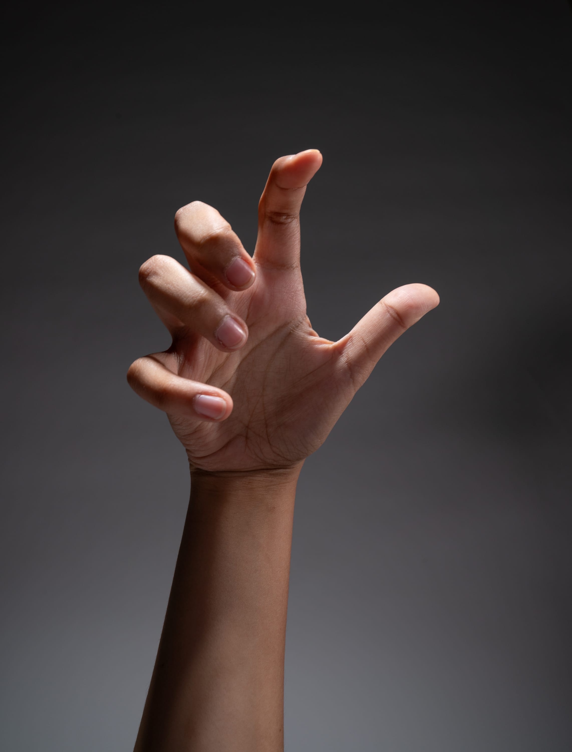 a single hand is outstretched against a dark gray background. the hand is lit dramatically from above, casting shadows and highlighting the tension in the fingers. the fingers are curled inward, creating a clawlike shape.