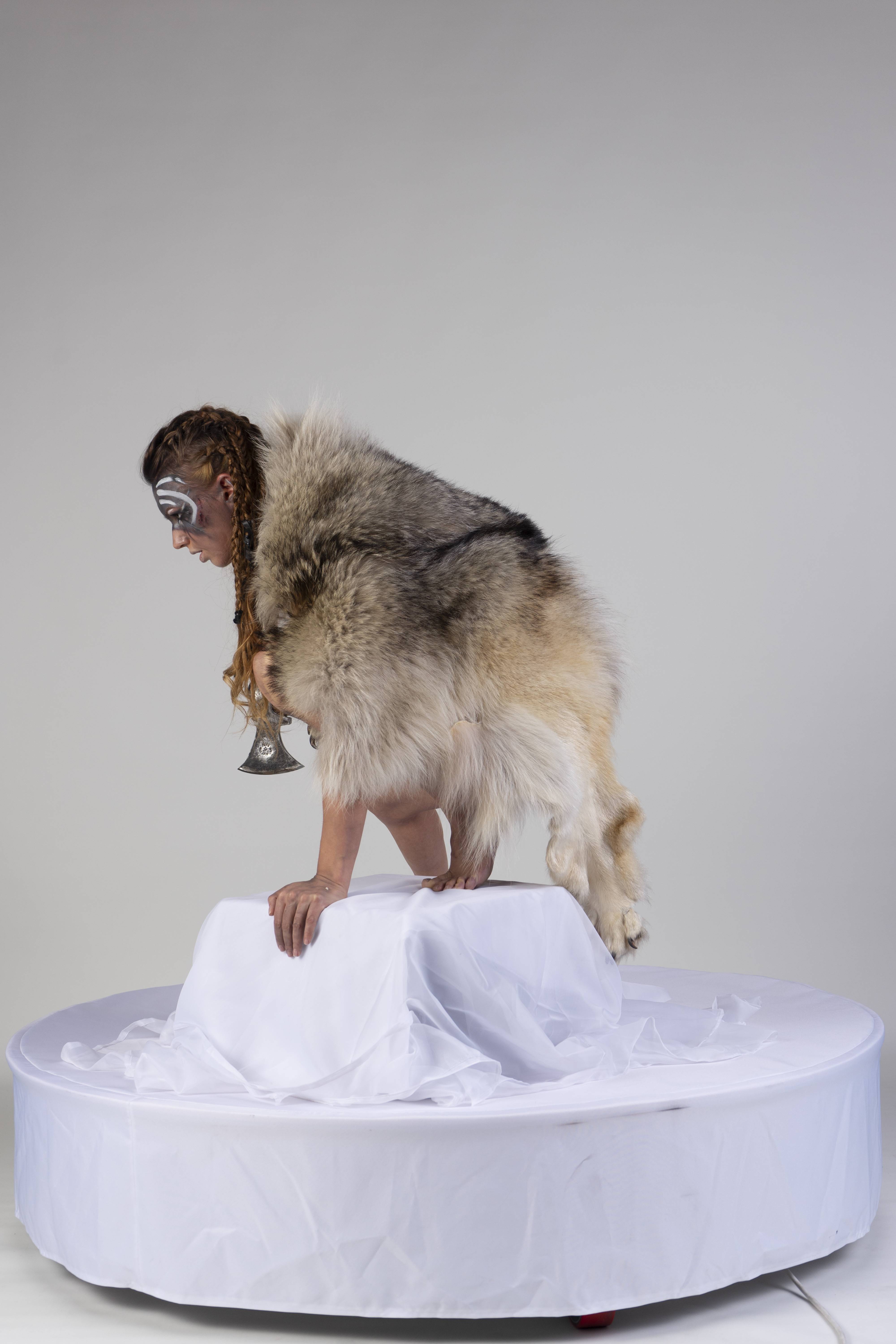 a woman dressed in a costume that includes animal fur and face paint is crouching on a white platform. she has braids in her hair and is holding an axe. the platform is covered in white fabric, and the background is a plain white.