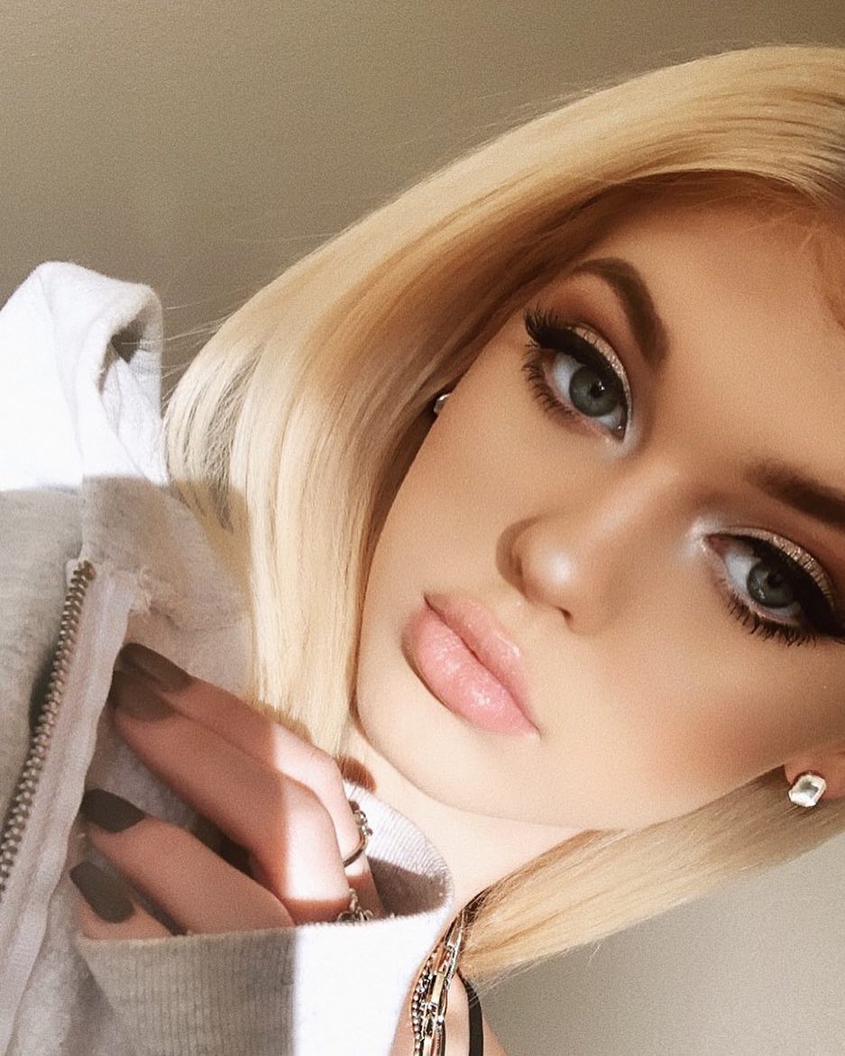 a closeup selfie of a woman with blonde hair, wearing makeup, and a gray hoodie. she has eyeshadow and lip gloss on. she also has jewelry.