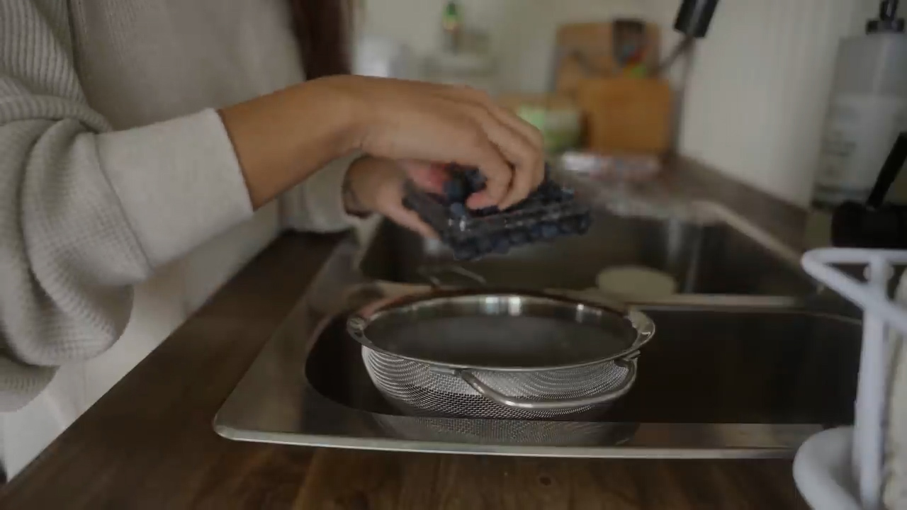 a woman is washing blueberries in a strainer over a kitchen sink. she is holding a plastic container of blueberries in her left hand and is using her right hand to rinse the blueberries.  the kitchen has wooden cabinets and countertops.