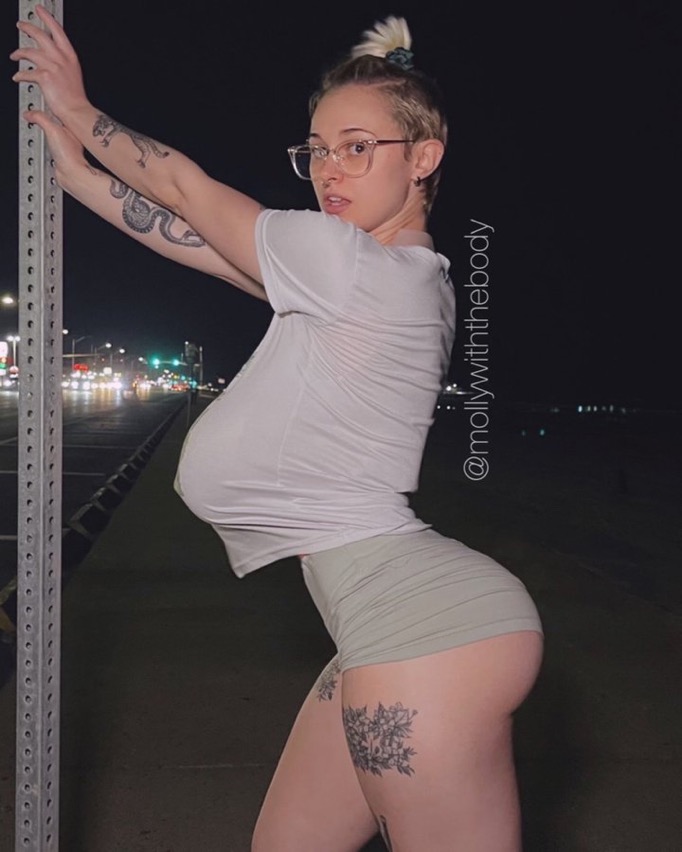 the image shows a young woman with short hair in a bun, wearing a white tshirt and light green shorts, posing outdoors at night in an urban setting. she is leaning against a metal pole and has several tattoos visible, including a snake and floral designs on her legs. she is wearing eyeglasses and appears to be posing for a photo. the overall impression is one of a fashion or social media post highlighting the woman's physique.  the watermark "@mollywiththebody" is visible on the image.