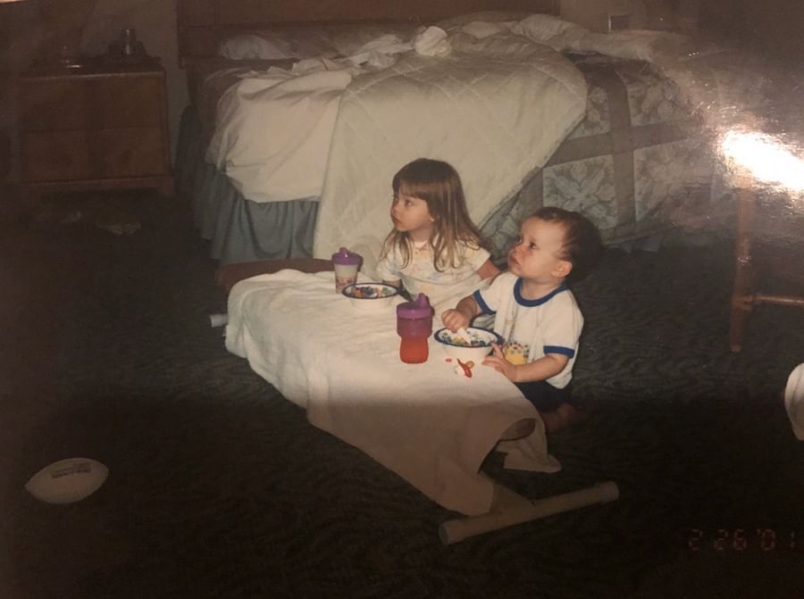 a slightly blurry photo of two young children, a girl and a boy who appear to be siblings, sitting at a makeshift table in what seems to be a hotel room.  they are eating cereal from bowls with spoons. the girl is slightly older and is sitting next to the boy.  the bed is visible in the background. the photo has the look of an old photograph, with a slightly faded and washedout quality. there are also some objects on the floor near the children, one of which is a small cup or bowl.