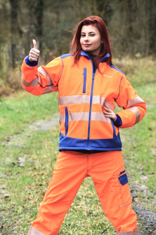 a redhead woman in an orange highvisibility safety jacket and matching pants stands outdoors in a grassy area, giving a thumbsup gesture. she appears confident and happy. the setting appears to be a natural environment, possibly a forest or park.  her clothing is workwearappropriate, suggesting a profession in a field requiring highvisibility apparel.