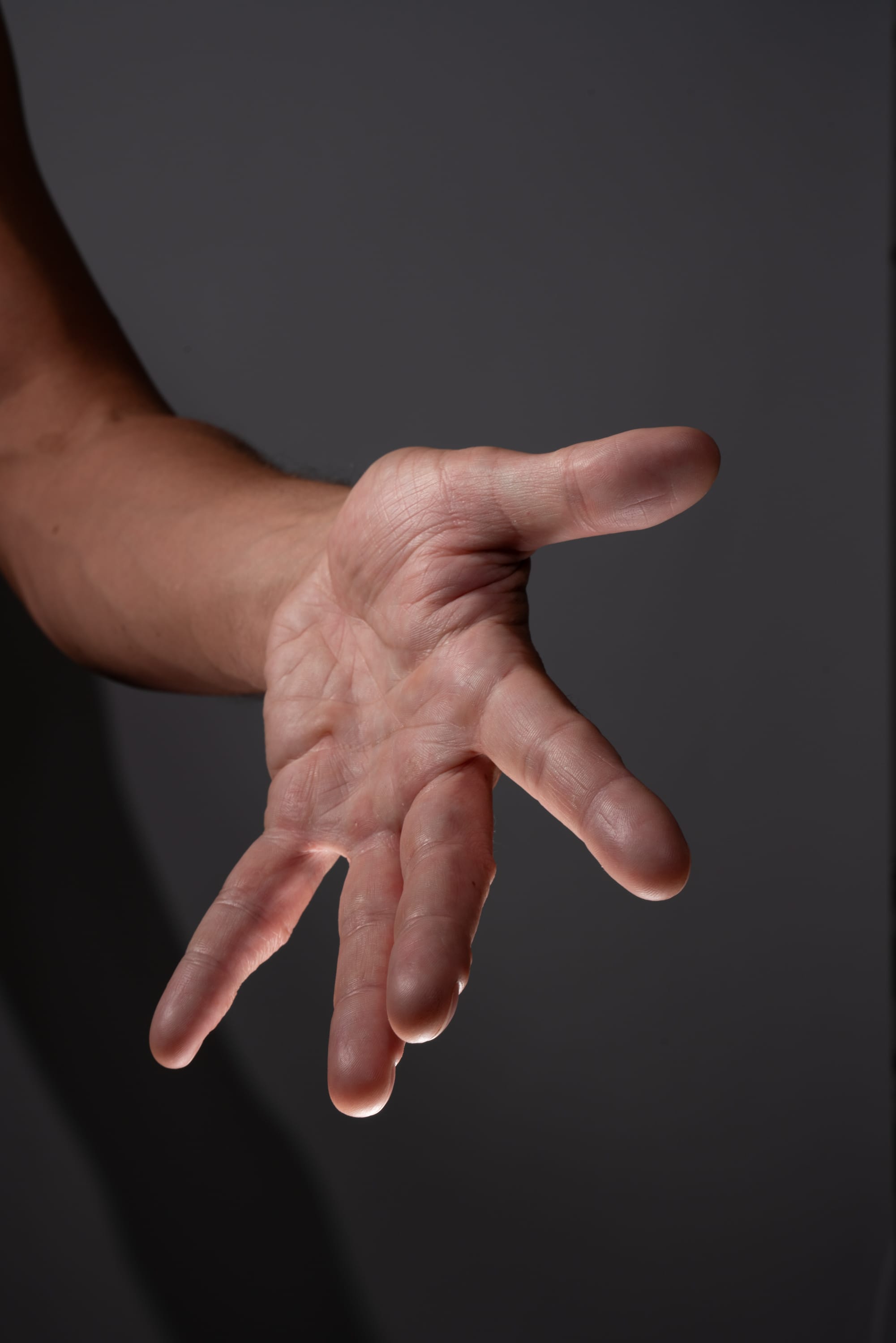 the image shows a man's open hand reaching forward against a dark gray background. the hand is illuminated by studio lighting, highlighting the texture and lines of the skin. the gesture suggests reaching out, asking for something, or offering assistance.