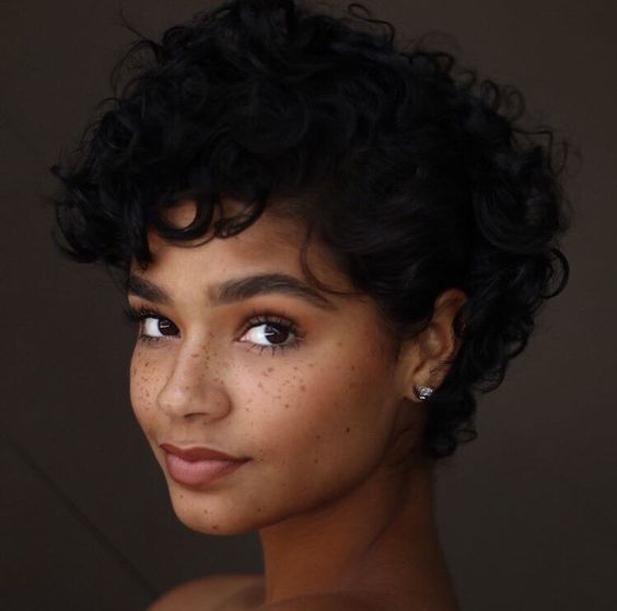 a closeup portrait of a woman with dark, curly, short hair. she has freckles on her face, wears small earrings, and looks directly at the camera with brown eyes. she has on makeup and her skin has a dark tone. the background is dark and blurred.