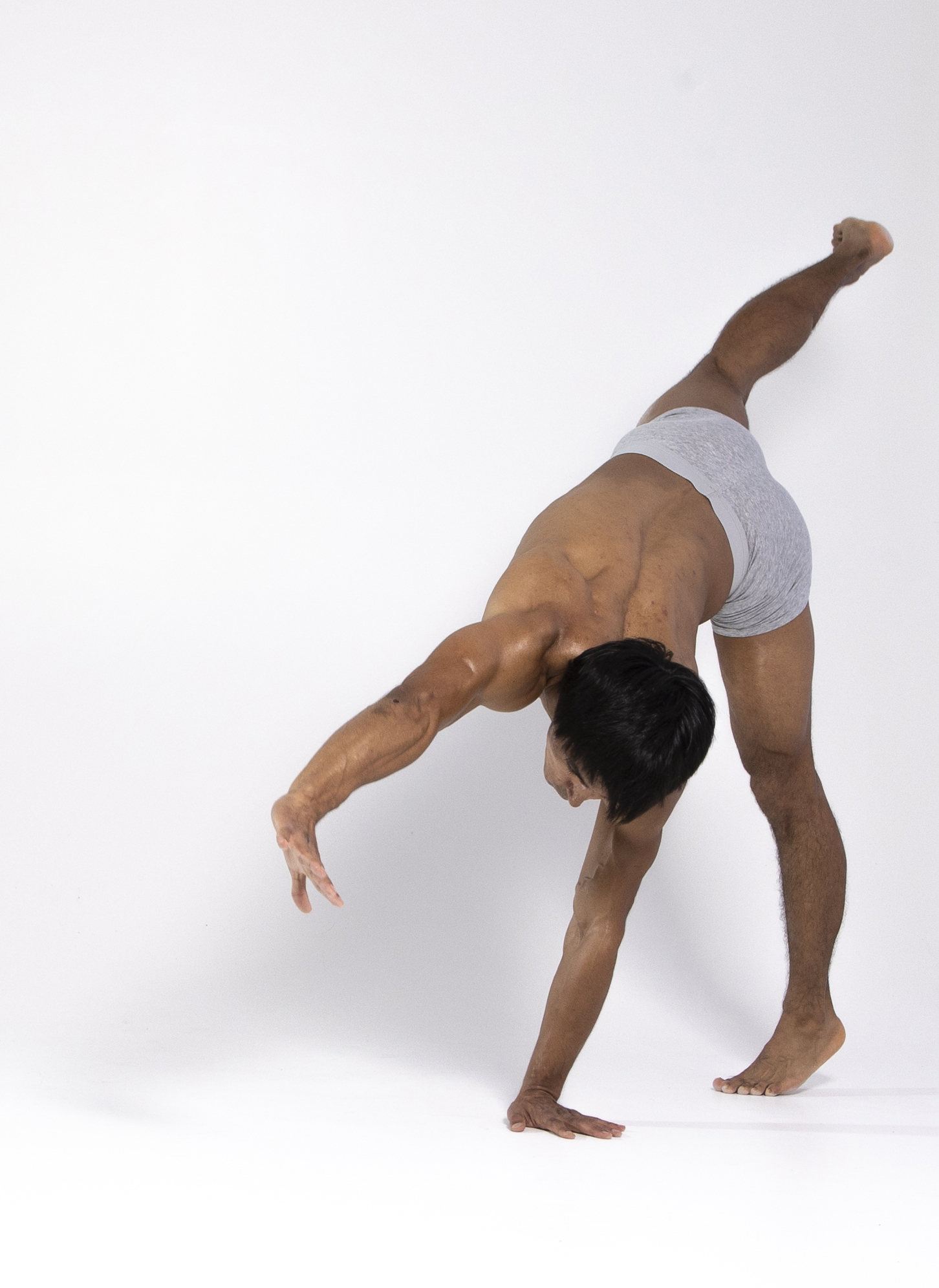a muscular man, wearing only grey underwear, is performing a contortion exercise in a studio setting. he is barechested, with one arm extended out to the side, and the opposite leg raised in the air. the photo is taken indoors against a plain white background.
