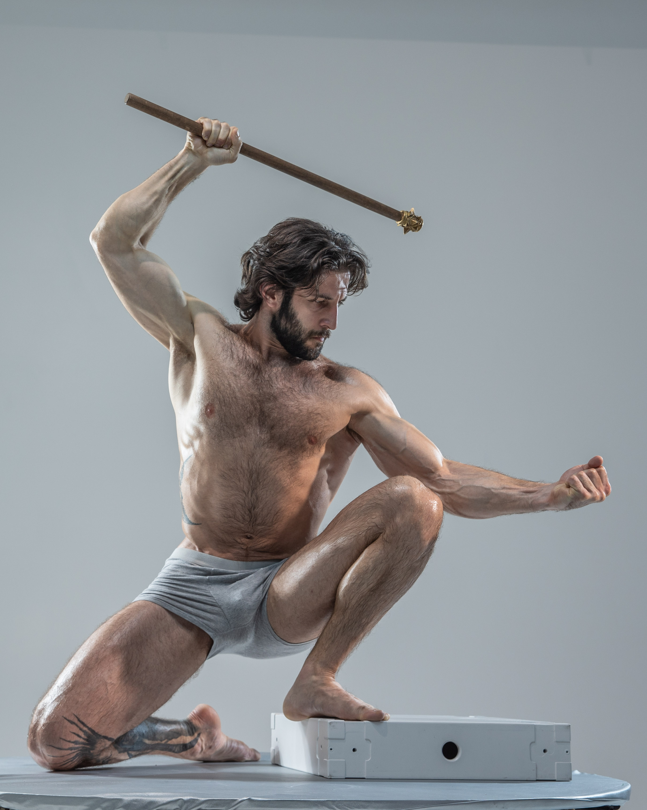 a muscular, hairy man in gray underwear holds a wooden staff above his head, captured midpose against a light gray studio backdrop. the lighting is soft, highlighting his physique and creating a dynamic, artistic image. his pose suggests strength and power.
