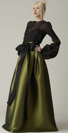 a woman in a long black and green dress poses in a formal portrait. she is wearing a black top with puffy sleeves and a matching belt. her skirt is a deep, rich green, and it has a slight shine to it. she is standing with her hand on her hip, and she is looking to the side. the background is a plain white wall.