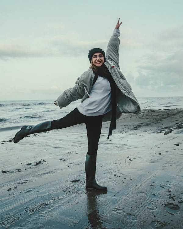 a young woman, wearing a grey puffy jacket, black beanie, and tall black boots, is joyfully leaping on a wet, sandy beach with one arm raised in the air.  the ocean stretches out behind her under a cloudy sky.  she is smiling broadly, showcasing a feeling of happiness and freedom. the overall mood is cheerful and energetic.