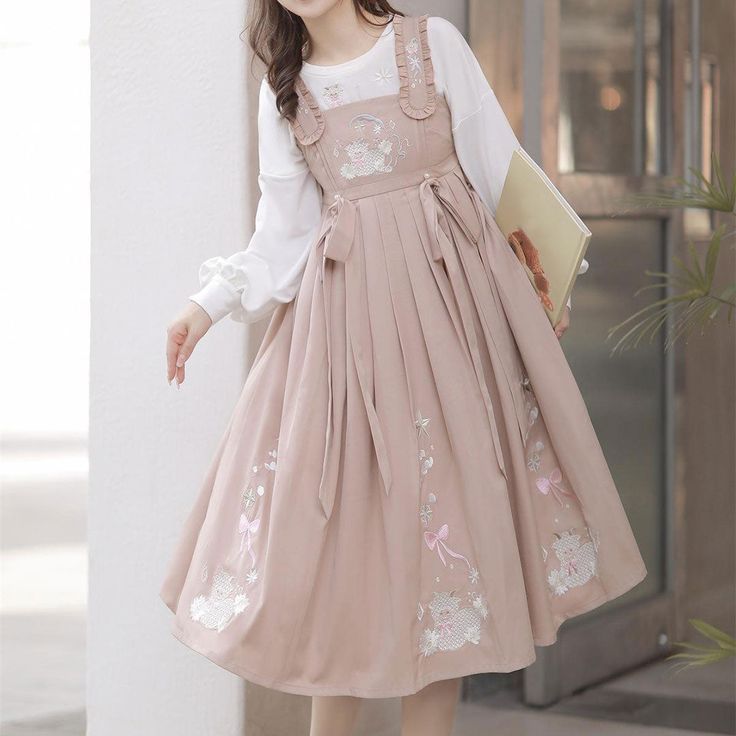 a young woman is wearing a pink dress with white embroidery. the dress is a lolita style and is very flowy. she is also wearing a white longsleeved shirt underneath and is holding a book in her left hand. the background is a nondescript building entrance, suggesting she may be at school.  the overall aesthetic is very soft and feminine.
