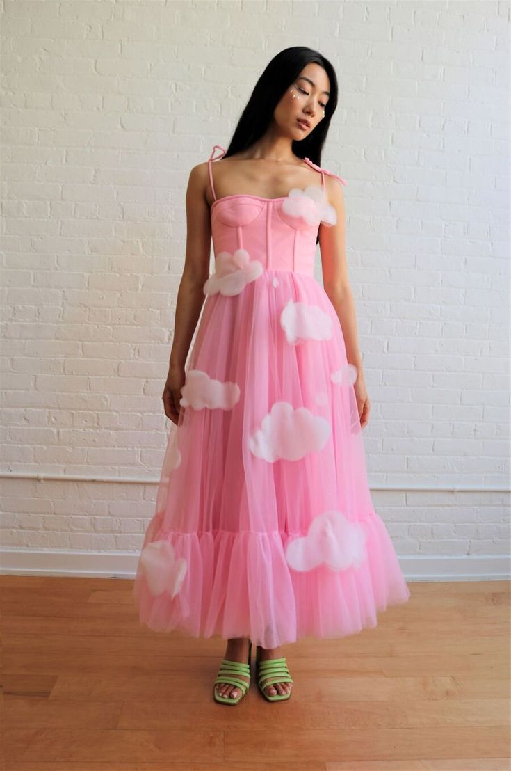 a woman in a pink tulle dress with cloudshaped embellishments. the dress is cinched at the waist and has a full skirt. the model is wearing green sandals and has her hair down. she is standing on a wooden floor in front of a white brick wall. the dress is very whimsical and fun, and the model looks confident and happy.