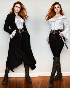 a woman poses for the camera in a pirate costume. she is wearing black pants, knee high boots, a white blouse, and a black over coat. she has a sword strapped to her waist.  she is standing against a white wall with light wood floors.