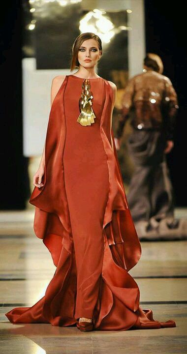 a woman in a beautiful orange dress with gold jewelry and matching high heels is walking down a runway. she is posing for the camera and looking very elegant and confident. the dress is very stylish and the woman's hair and makeup are done perfectly. she is a true fashion icon.
