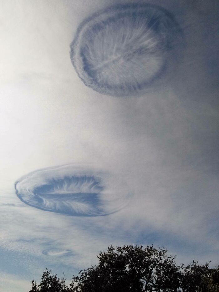 a photo of two lenticular clouds in a clear, blue sky. the clouds are white and have a distinct oval shape. the top cloud is larger and more prominent than the bottom cloud. there is some other cloud cover, but it is very thin. the sky is a light blue, and there is a hint of sunlight shining through. the clouds are a beautiful and unusual sight, and they add a touch of wonder to the sky.