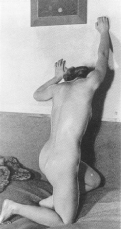 a black and white photograph of a nude male model posing in a studio. the model is facing away from the camera, with their arms raised above their head and their hands touching the wall. the model's body is visible from the back, showing the muscles and contours of their physique. the background is simple and uncluttered, focusing attention on the subject. the photograph has a vintage or antique quality to it, suggesting it may be a historical piece.