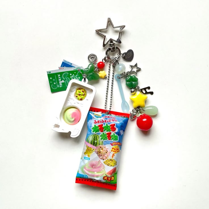 this image shows a keychain with various charms. the charms include miniatures of a toy food item resembling a candymaking set, a candy package, a star, a spoon, a fourleaf clover, and a watermelon. the keychain is adorned with beads and various small trinkets.