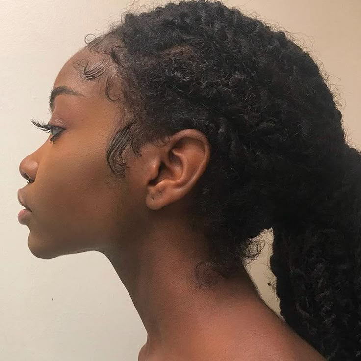 this is a side profile portrait of an africanamerican woman with natural, curly hair styled in a ponytail. her edges are styled and she has a nose piercing. she has makeup on and is facing left. the background is a lightcolored wall.