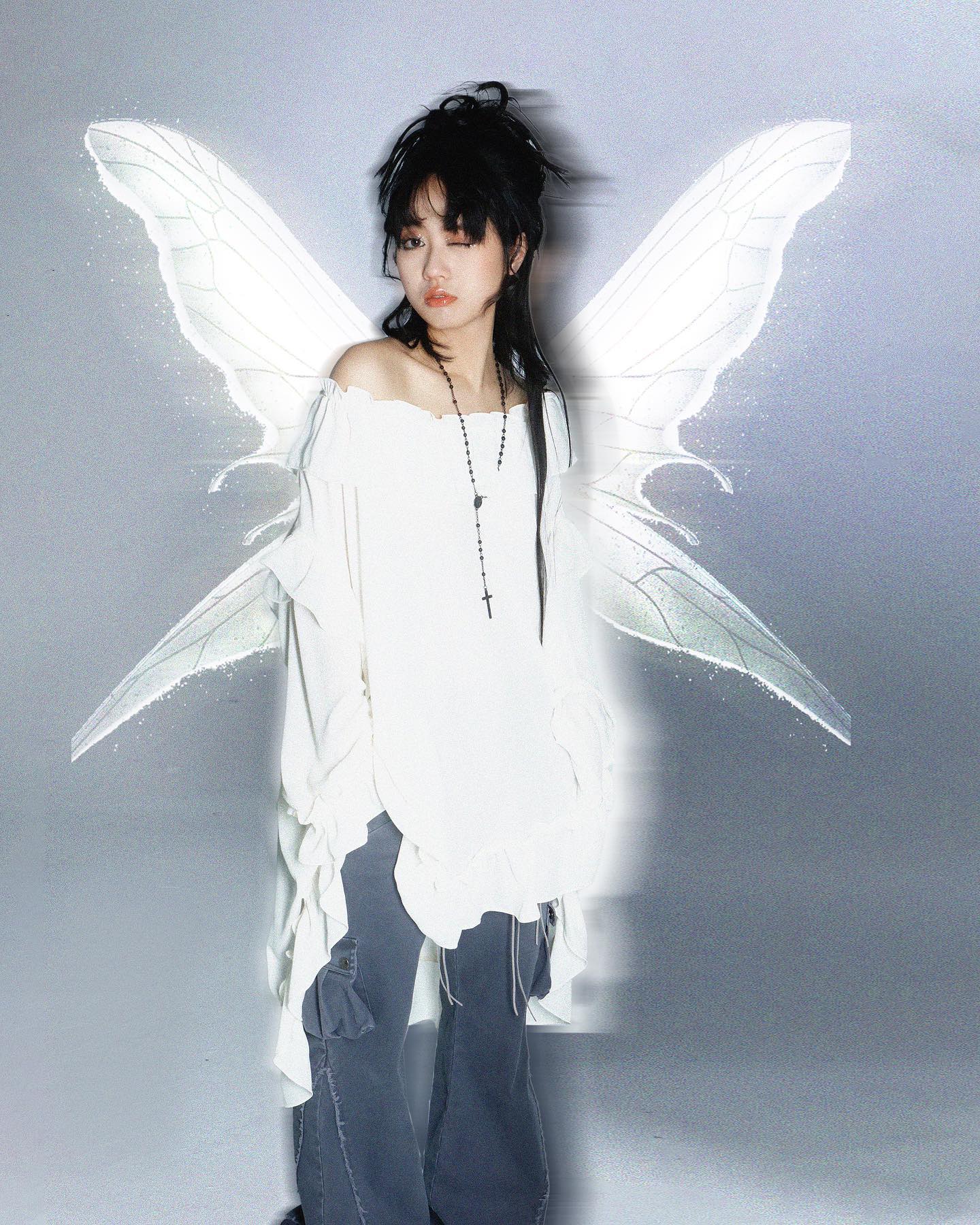 the image portrays a young asian woman, possibly a model, dressed in alternative fashion. she's wearing a voluminous, offshoulder white shirt with ruffles and grey cargo pants.  a large, delicate pair of translucent fairy wings are digitally added behind her, enhancing a dreamy and ethereal aesthetic.  she wears a simple black cross necklace. her dark hair is styled with bangs. the overall style is a blend of soft grunge, and ethereal fantasy. the background is a light grey, and the lighting is soft and diffused.  the photo has an artistic and moody feel, emphasizing the model's mysterious expression.