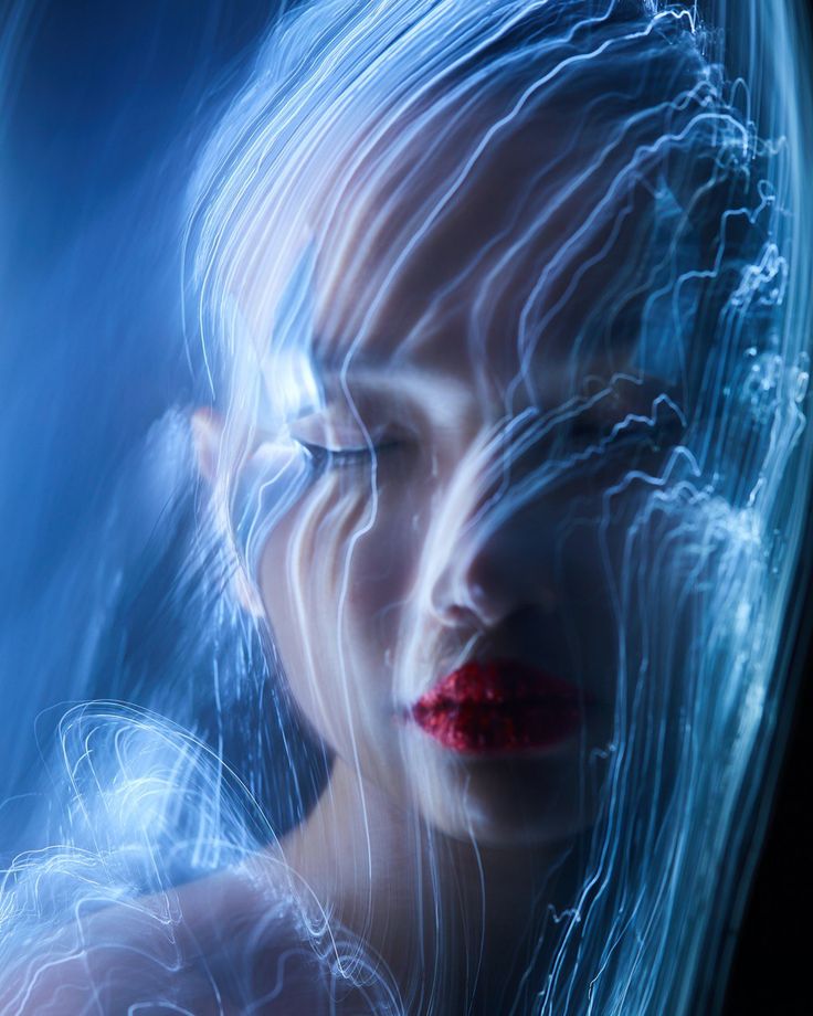the image is a stunning light painting portrait of a woman.  her face is partially obscured and blended with streaks of vibrant blue light, creating an ethereal and surreal effect. the long exposure photography technique results in dynamic light trails that seem to flow around her head and face. her red lipstick stands out in contrast to the cool blue tones. the overall mood is mysterious and artistic, suggesting a blend of fantasy and reality.
