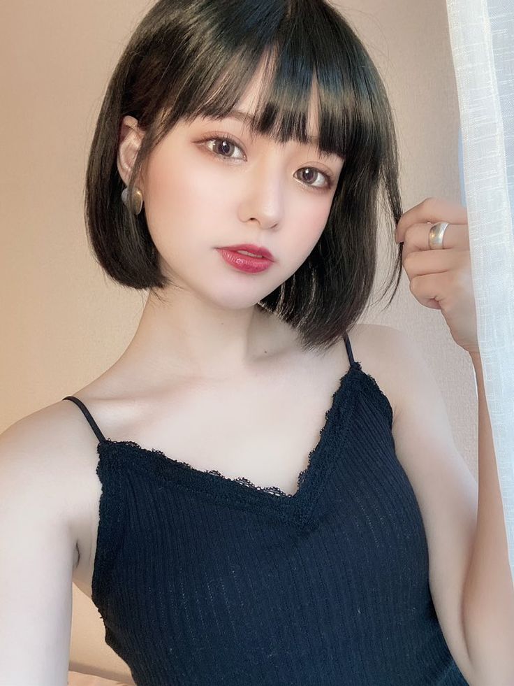 a young woman with short black hair and bangs is looking directly at the camera with a soft smile. she is wearing a black tank top with lace trim. she is posing with her right hand resting on the left side of her face.