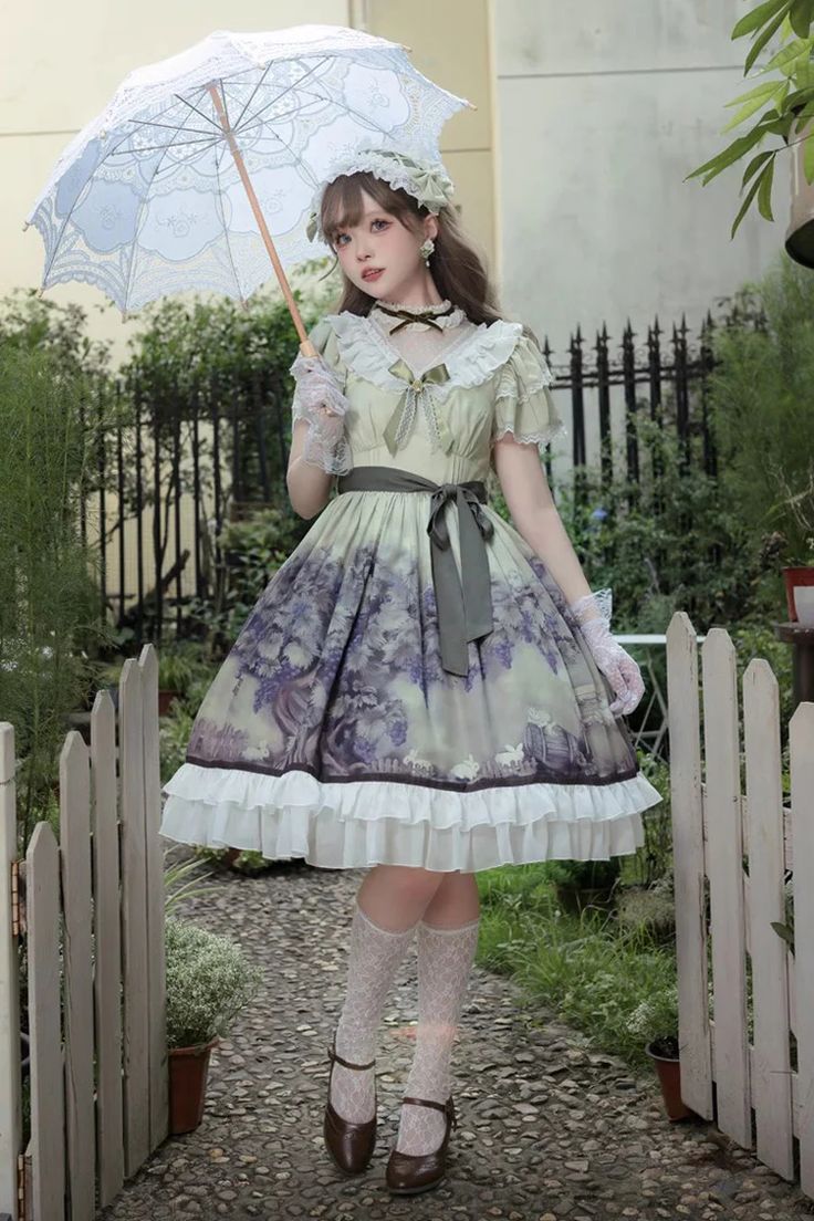 a woman in a flowing green, white, and purple lolita fashion dress stands on a stone path in a garden. she holds a white lace parasol and is smiling subtly at the camera. the dress features a print of trees and a forest and has multiple layers of fabric and lace detailing. she wears brown mary jane shoes and white socks. the background features green plants and a white picket fence. the lighting is natural and bright, with a slightly overcast sky.