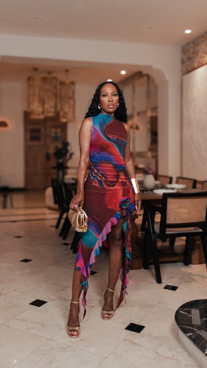 a fullbody shot of a stylish africanamerican woman in a vibrant, multicolored dress. she's posing elegantly in a beautifully decorated dining room.  the dress has a high neck and a ruffled, asymmetrical hem. she's carrying a gold clutch and wearing rose gold highheeled sandals. the setting is luxurious, with marble flooring and upscale furniture.  the lighting highlights her pose and the details of the dress.