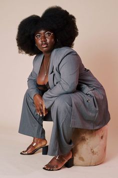 a black woman with a large afro is wearing a grey suit and matching pants, black heels and a pair of trendy gold rimmed glasses. she's sitting on a wooden stool and looking to the side. the background is soft cream with soft light illuminating the scene.  the woman is posed in a relaxed yet stylish way, her hair styled elegantly. her makeup is subtle, but it highlights her features. the image captures a confident and stylish woman.