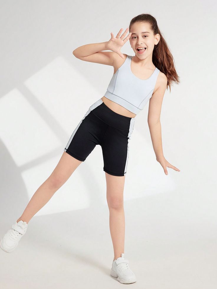 a young girl, likely a tween or preteen, is dynamically posing in a light blue sports bra and black biker shorts with white stripes.  she's wearing white athletic shoes and has her hair pulled back. she's smiling and playfully waving her hand to the side, suggesting energy and activity. the setting is a bright, clean white studio background. the overall impression is one of youth, health, and active living, with a focus on comfortable, stylish sportswear.