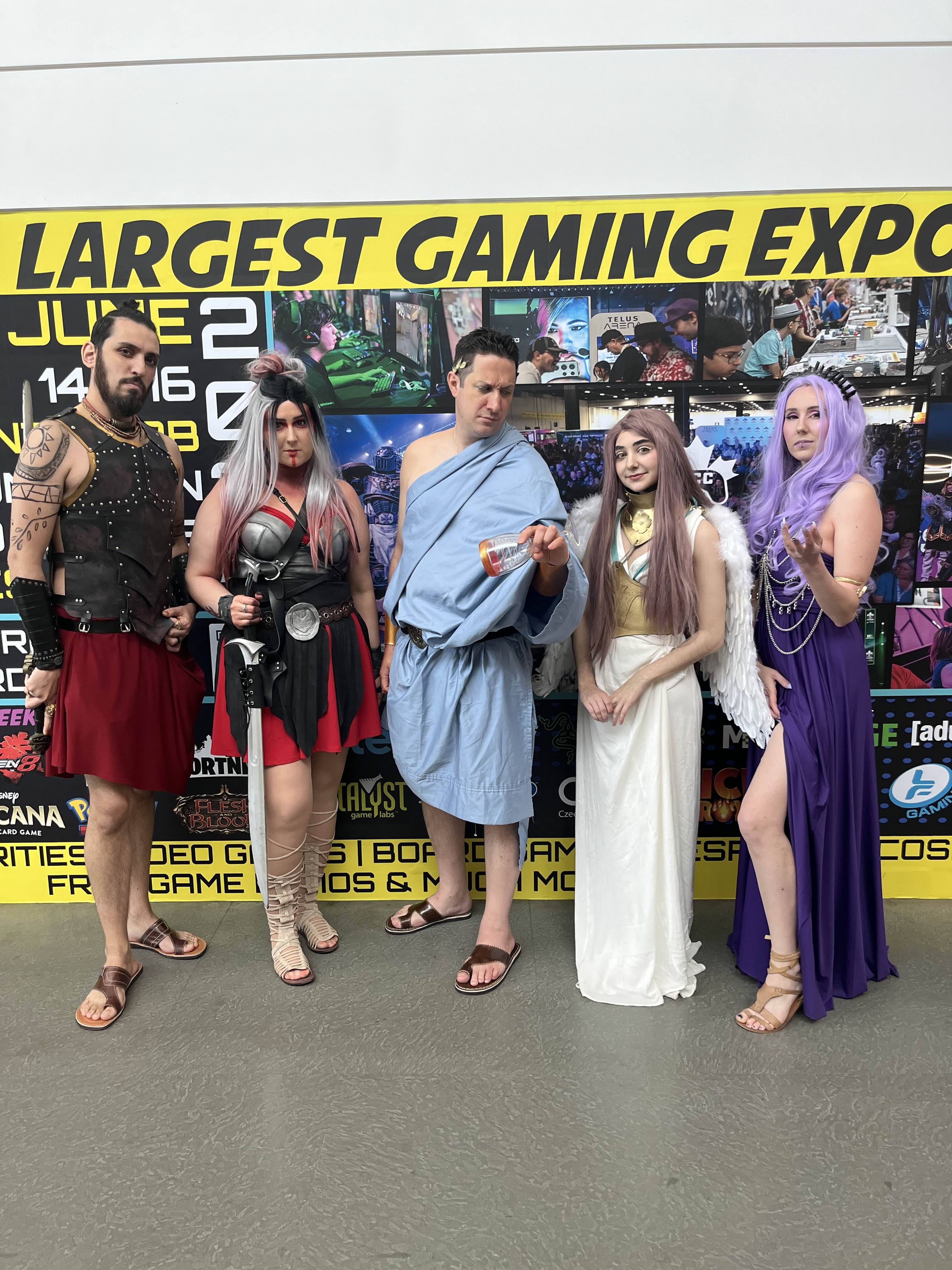 group of five cosplayers pose in front of a banner for a gaming expo.  they are dressed as various characters: a kratosinspired character, a female warrior, a male in a greek tunic (possibly representing a god), a persephonelike character, and a goddesslike character in purple.  the setting appears to be an event hall or convention center.  the banner prominently features the words "largest gaming expo".