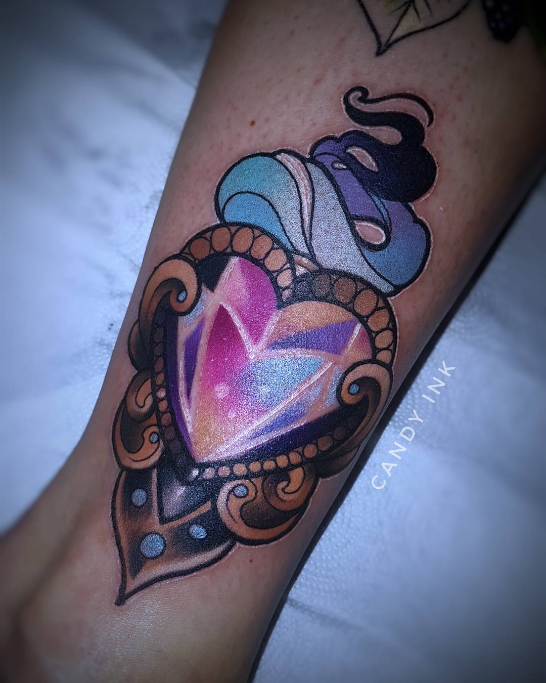 a closeup photograph showcasing a vibrant tattoo of an ornate heartshaped jewel nestled within an elaborate frame. the jewel shimmers with hues of pink, purple, and blue, while a swirling cloud in matching colors appears to emanate from its top. the tattoo is set against the backdrop of the recipient's arm.