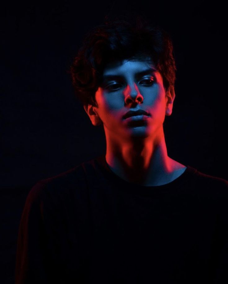 a young man with a serious expression on his face is shown in a portrait. the man is lit by red and blue light, casting dramatic shadows on his face and neck. he is looking directly at the camera, wearing a black shirt. his face and expression give the impression of someone who is thoughtful or introspective.  the image is dark and moody with a strong contrast between the light and shadow, which adds to the dramatic effect.