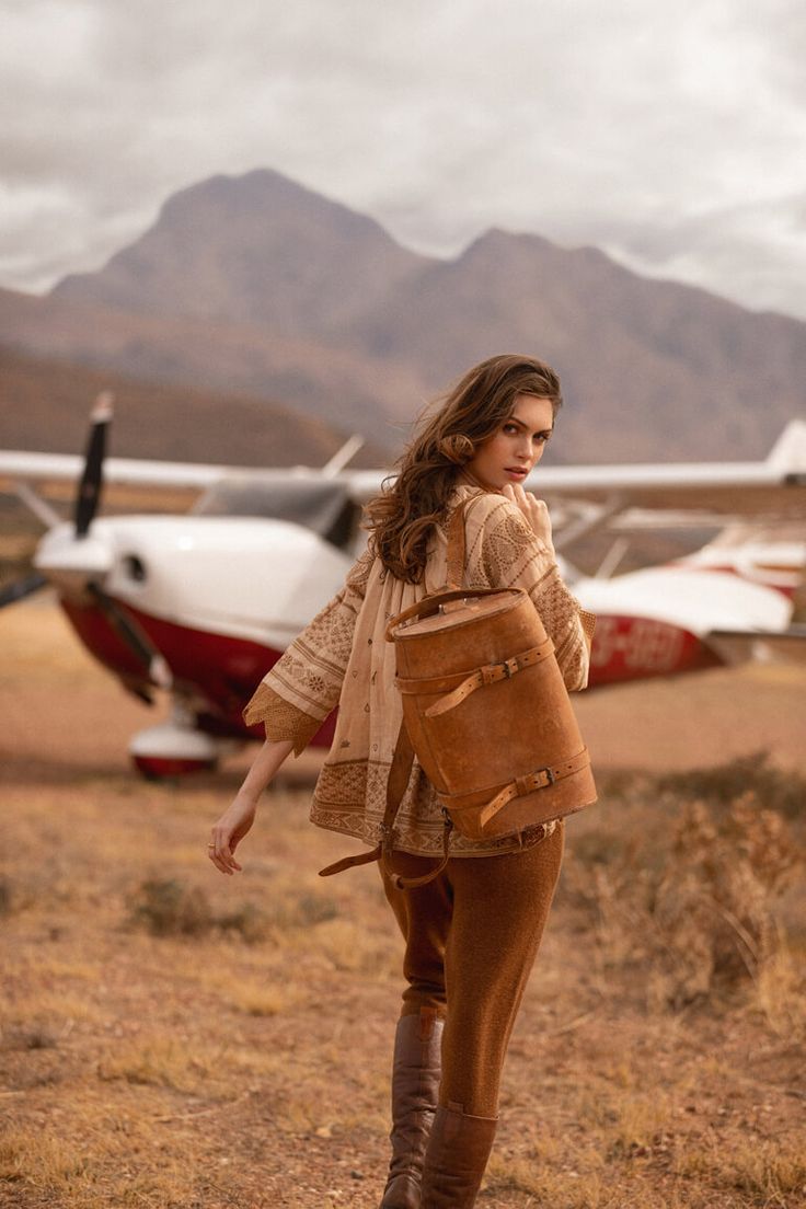 a full shot of a fashion model walking away from the camera. she is wearing a beige patterned blouse, brown corduroy pants, brown leather boots and a brown leather backpack. she is walking on a runway near a small airplane in front of a mountainous landscape. the overall tone of the image is warm and earthy, evoking a sense of travel and adventure.