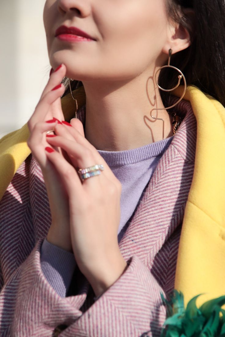 closeup portrait of a stylish woman wearing a yellow coat over a purple sweater.  she's wearing gold abstract earrings and a ring. her hands are gently clasped together, and her red manicure is visible. the image focuses on her clothing and accessories, highlighting the textures and colors. the overall mood is elegant and fashionable.