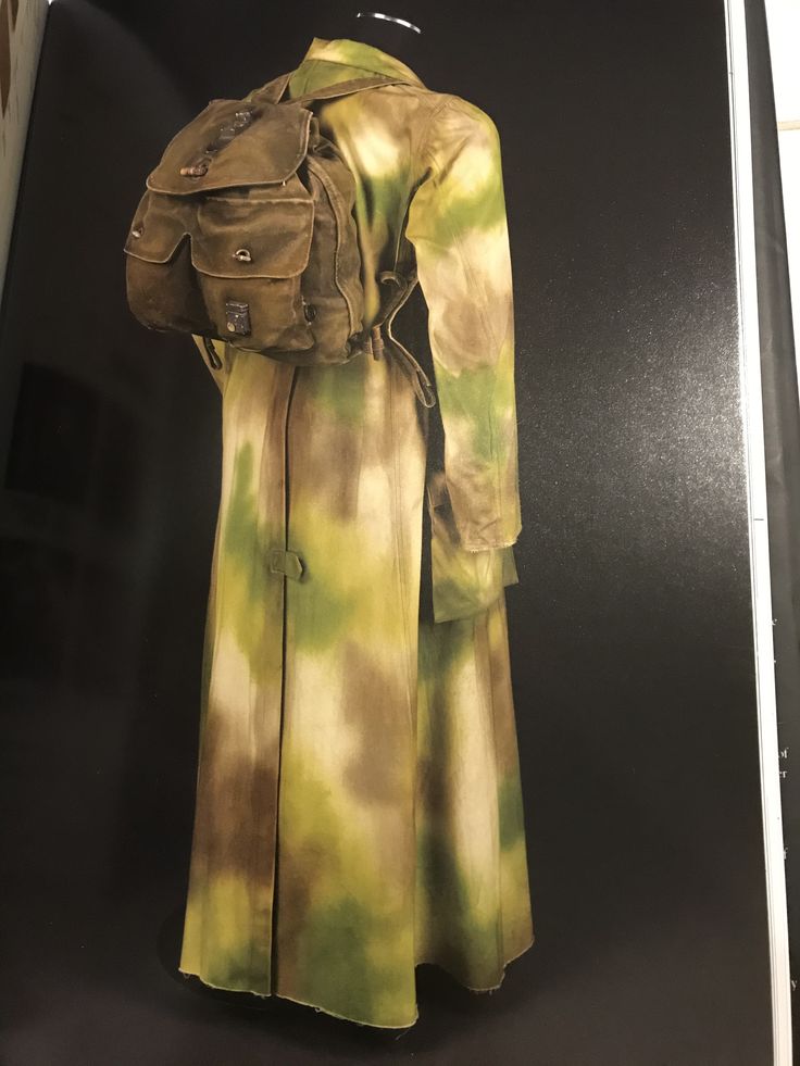 this image shows a closeup of a vintage military coat with a camouflage pattern. the coat is a long, flowing style, with a green, brown, and beige camouflage pattern. the coat is being displayed on a mannequin in a museum exhibit. the coat has a backpack attached to it.