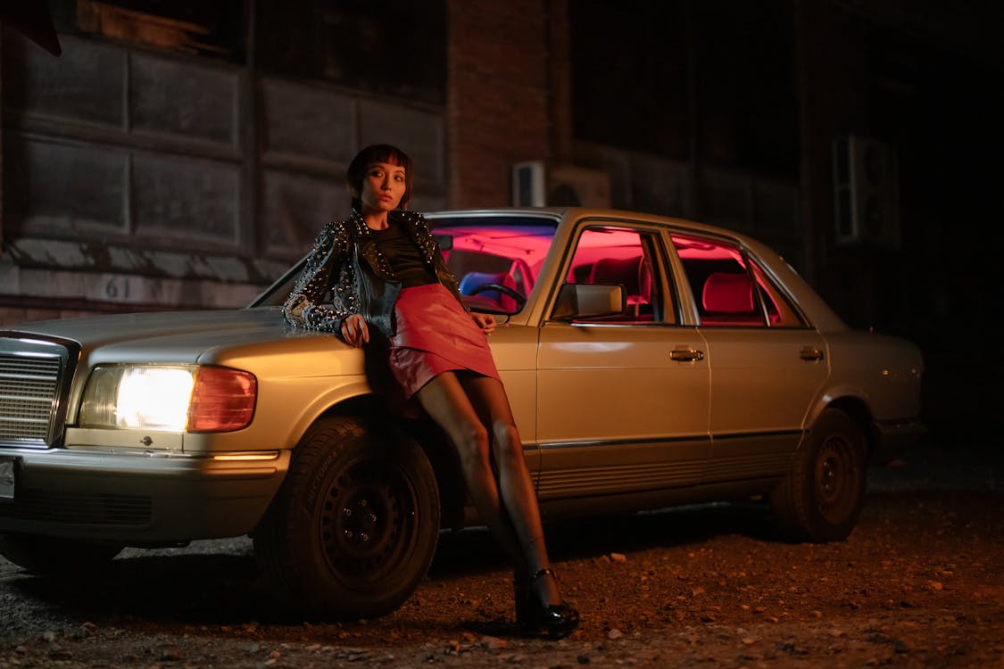 the image shows a stylish young woman with short dark hair, wearing a black leather jacket and a red mini skirt, posing confidently on the hood of a vintage beige mercedesbenz sedan. the car's interior is illuminated with pink and blue neon lights. the setting is a dark, urban street at night. the overall mood is retro and moody, with a focus on fashion and attitude.  the woman's pose and the car's aesthetics evoke a feeling of rebellion and coolness.