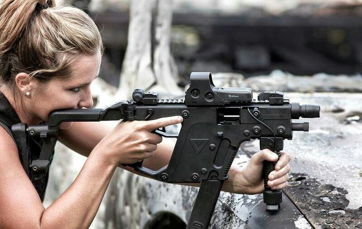 a woman is holding a vector smg with an optic sight and a vertical foregrip. she is biting the gun's stock. the background appears to be a damaged or destroyed building.