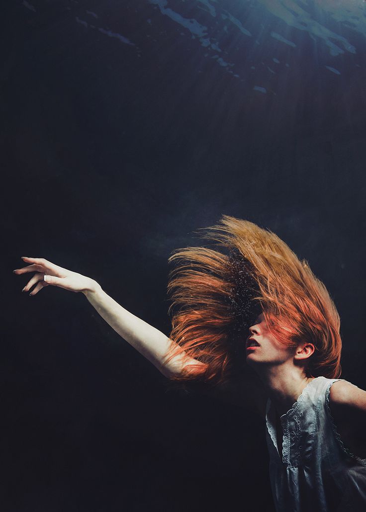 a woman with long red hair is submerged underwater, her hair flowing around her face. she is wearing a white dress and has one arm outstretched, reaching towards the surface. the image is dark and mysterious, giving a sense of dreamlike beauty and ethereal quality.