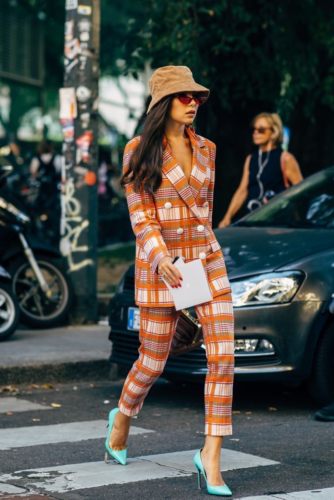 a stylish woman walks across a city street. she's wearing a vibrant orange and white plaid pantsuit, a beige bucket hat, and mint green high heels.  she carries a small white envelope and a brown handbag.  the background includes a car and a motorcycle. the overall aesthetic is trendy and sophisticated street style.