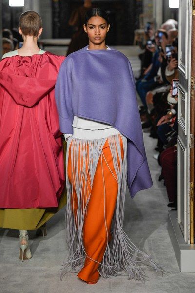 two fashion models walk down a runway in front of a seated audience. the model on the left is wearing a red cape with a green underskirt and grey shoes. the model on the right is wearing an orange skirt with a long grey fringe, a white shirt, and a purple cape. the model has her dark hair pulled back from her face and she is wearing a silver necklace.
