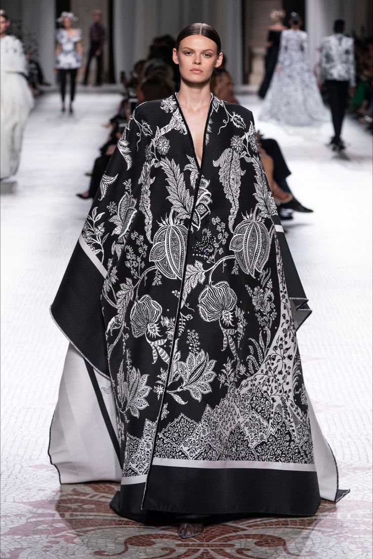 a model walks the runway at a fashion show wearing a long, black and white capestyle gown. the gown features an intricate floral pattern in shades of black and white, with a long train and capelike sleeves. the model's hair is neatly pulled back, and her makeup is simple and elegant. the background is blurred, showing other models and audience members. the overall impression is one of high fashion and elegance.