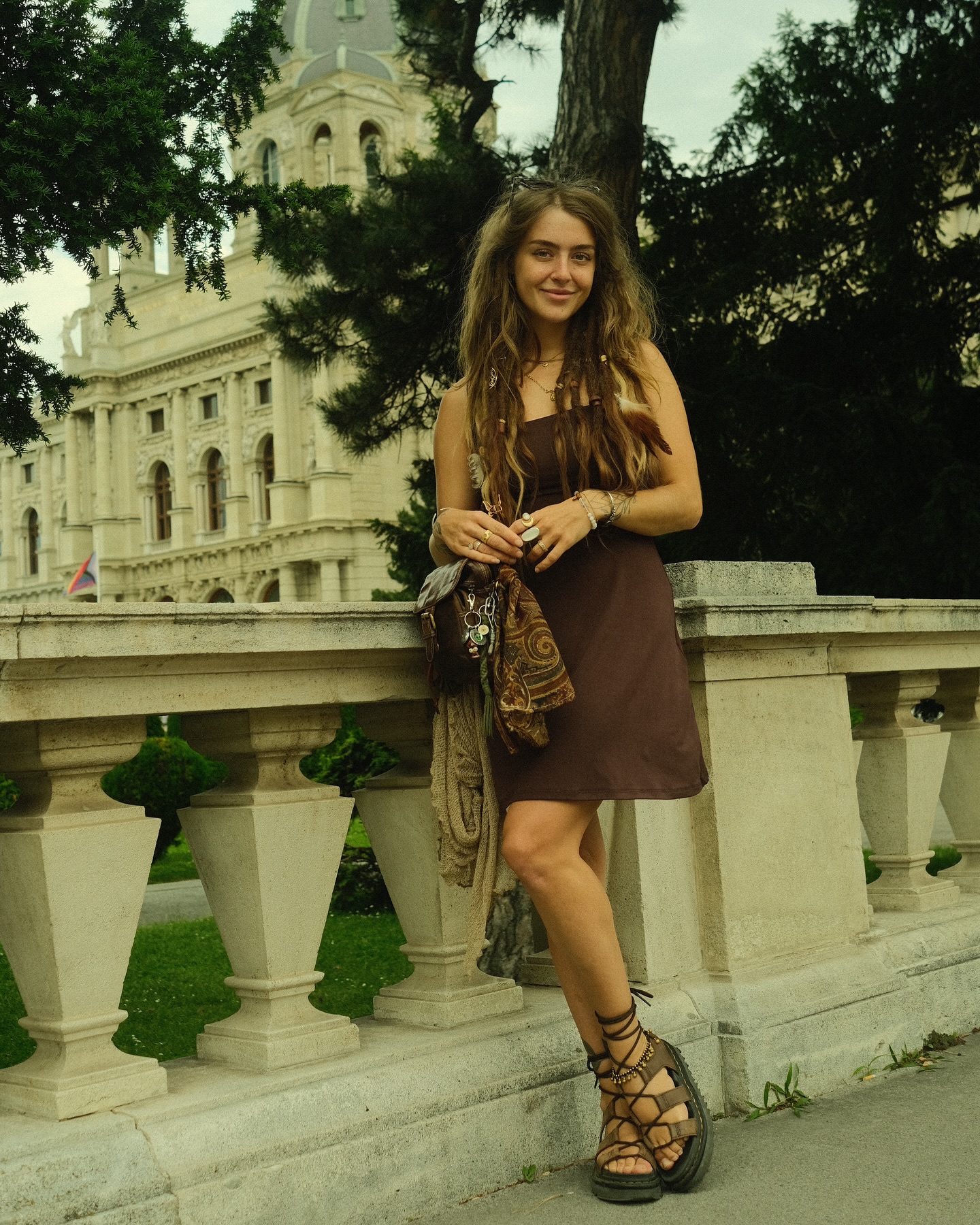 a young woman with long brown dreadlocks and a bohemian style stands leaning against a stone balustrade in front of a large building. she is wearing a brown sleeveless dress, brown sandals with laces, and several pieces of jewelry. she carries a paisley patterned scarf and a small bag. the building behind her appears to be a museum or historical building with classical architecture.  the setting is outdoors, in a parklike area. the overall aesthetic suggests a travel or tourism context. the image is somewhat nostalgic or vintage in its coloring and quality.