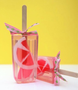 two homemade popsicles on a yellow background. the popsicles are made with grapefruit, and are wrapped in pink ribbon with green polka dots. the sticks are wooden.  the popsicles are sitting on a white surface.