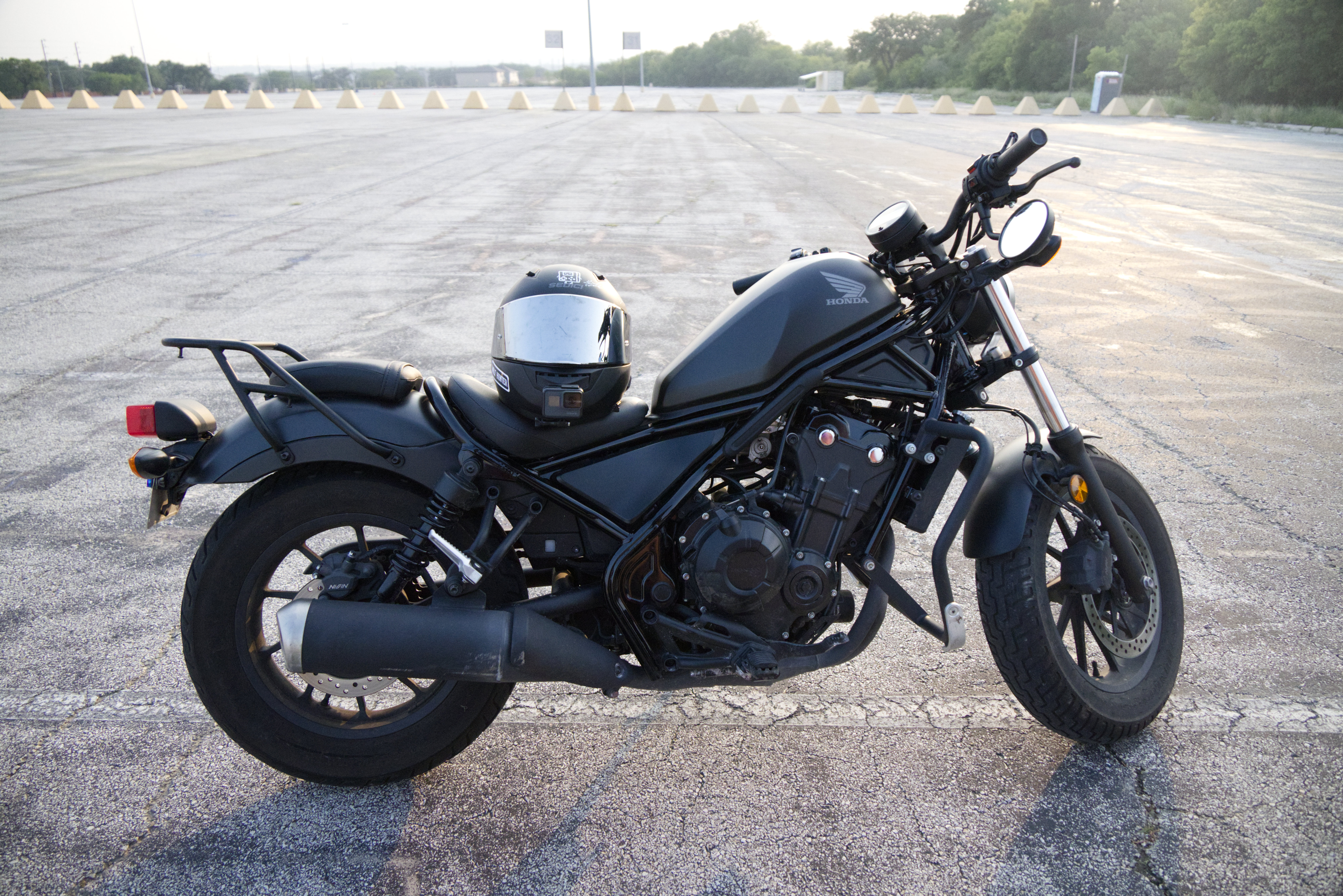 a black honda cmx500 rebel motorcycle is parked in an empty parking lot with a black fullface helmet resting on the seat. the sun is shining, and the asphalt is visible. the motorcycle is in good condition and appears to be new or wellmaintained. the image is taken from a slightly elevated angle, offering a view of the motorcycle from the side.  the background shows a flat, empty parking area with evenly spaced traffic cones in the far distance.