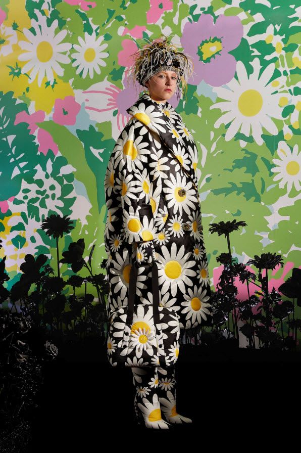 a fashion model is posing in a black and white daisy print coat, matching bag, and boots. the model is standing against a colorful floral backdrop. the clothing is highfashion and is a chic and stylish example of contemporary fashion.