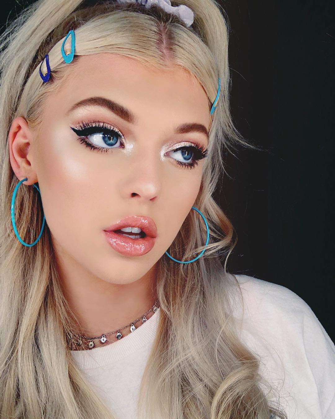 a closeup image of a woman with blonde hair styled in a halfup ponytail with blue hair clips. she has blue eyes and is wearing heavy makeup, including eyeliner, highlighter, and glossy lipstick. she is wearing a necklace and matching blue hoop earrings. she is wearing a white tshirt and looking off to the side.