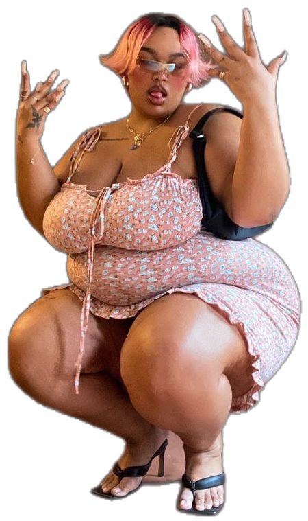 a plussize woman with pink hair and pink sunglasses is crouching while wearing a floral dress and sandals. she has jewelry on and tattoos on her arms.