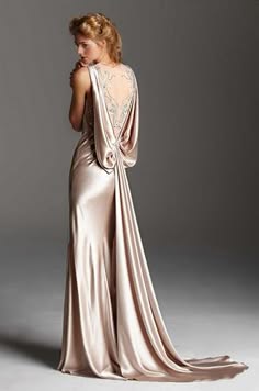 a beautiful woman in a luxurious, flowing silk dress. she is wearing a beige, floorlength gown with a cowl neckline and an open back. the fabric drapes elegantly around her body, creating a very glamorous and sophisticated look.  the dress features a delicate beading detail around the neckline and bodice. the model is posing with a hand on her hip and a thoughtful expression on her face. the background is a simple grey studio backdrop.