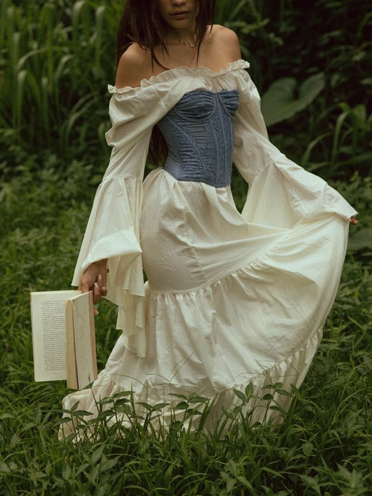a woman stands in a lush green garden wearing a long, white, offtheshoulder dress with a blue corsetstyle top. she holds a book in her left hand, and the dress has ruffled details around the neckline and hem. the long sleeves of the dress are wide and flowing. the garden is filled with tall grass and other plants.